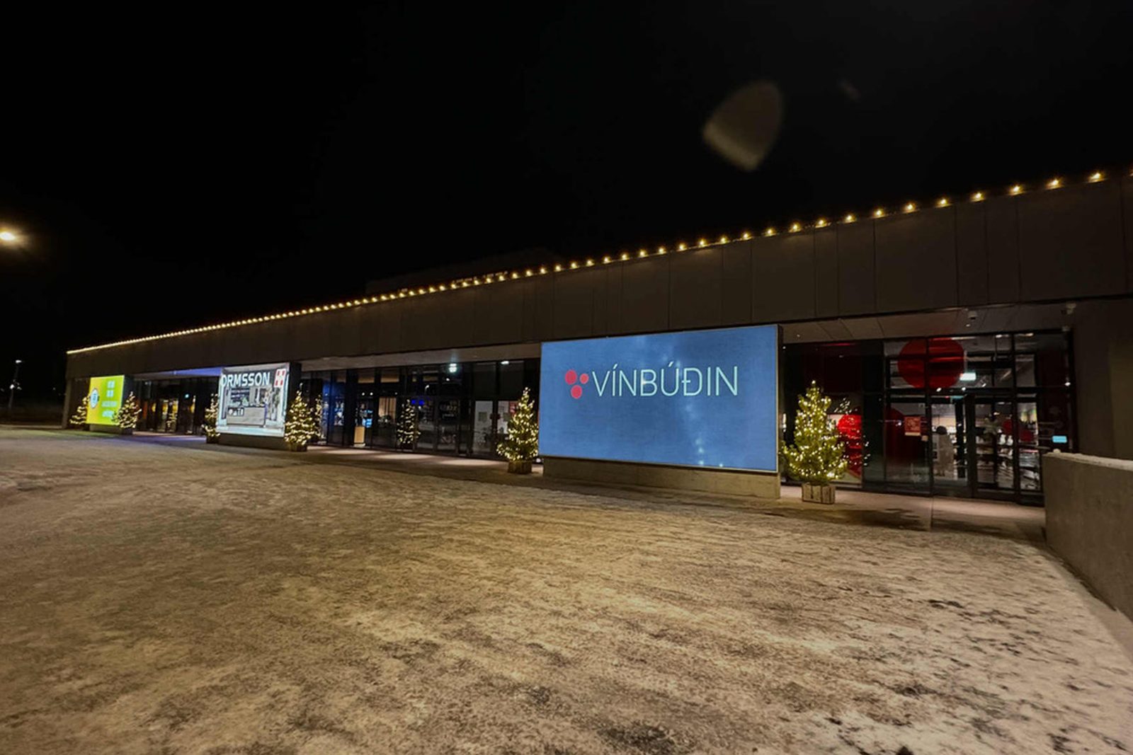Verslunarkjarni að kvöldi með upplýstum þakkanti og jólatrjám við inngangana