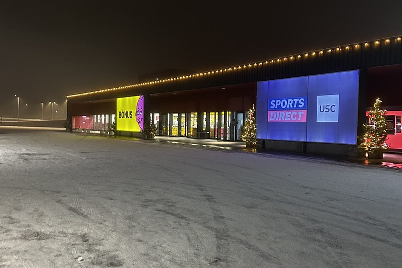 Verslunarhús í snjó með ljósum eftir þakkantinum og upplýstum trjám við innganga
